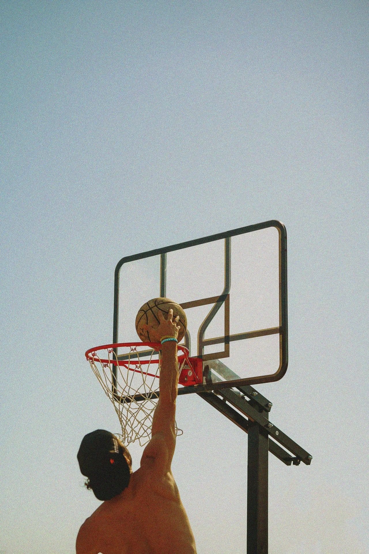 A shirtless person in a backward cap jumps to dunk a basketball into an outdoor hoop against a bright, clear blue sky. The image is shot from below, capturing the action and the ball just above the rim.