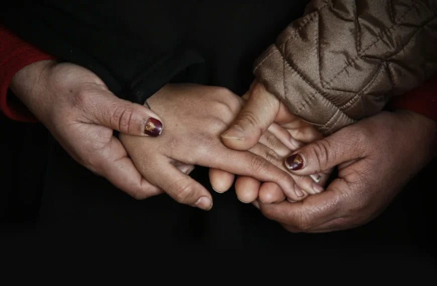 Three hands are shown: an adult’s hand holding a child’s smaller hand from above, while another adult hand supports from below. The adults have painted nails and wear a red sleeve and a brown quilted jacket. The background is dark, highlighting the hands in a gesture of care.