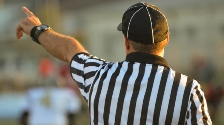 A football referee is seen from behind, wearing a black and white striped shirt and black cap, raising his left arm and pointing left, likely signaling a call on the field in accordance with national football league rules, with blurred players and background.