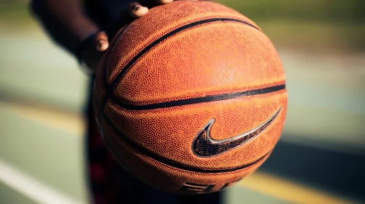 A close-up of a person holding a textured orange basketball with a black Nike logo. The focus is on the ball, inviting viewers to reflect: are sports fans less intelligent, or simply passionate? The lighting highlights the basketball’s detailed surface.