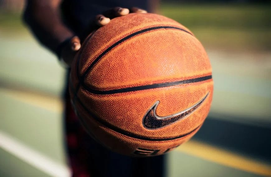 A close-up of a person holding a textured orange basketball with a black Nike logo. The focus is on the ball, inviting viewers to reflect: are sports fans less intelligent, or simply passionate? The lighting highlights the basketball’s detailed surface.