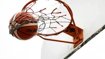 A basketball passes through an orange hoop and net attached to a white backboard, captured from below against a plain white sky. The ball is halfway through the net, suggesting a successful shot.