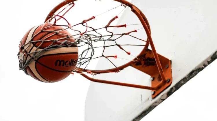 A basketball passes through an orange hoop and net attached to a white backboard, captured from below against a plain white sky. The ball is halfway through the net, suggesting a successful shot.