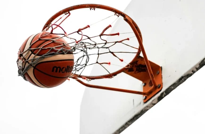 A basketball passes through an orange hoop and net attached to a white backboard, captured from below against a plain white sky. The ball is halfway through the net, suggesting a successful shot.