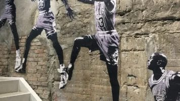 Black-and-white mural of a basketball player in a Bulls jersey performing a slam dunk decorates the rough concrete wall above a stairwell, capturing multiple dynamic poses that evoke the golden era of basketball as you climb the stairs.