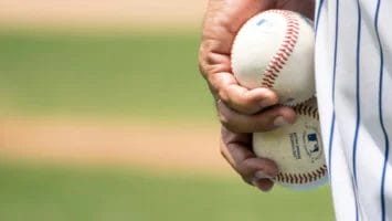 A close-up of a hand holding two white baseballs with red stitching captures the spirit of the New Era of MLB Analysis. The person wears a striped jersey and black wristband, set against a blurred baseball field with green grass and dirt.