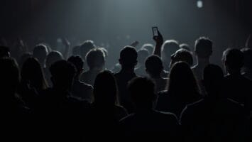 A crowd of people stands in a dimly lit venue facing a bright spotlight. Most faces are in shadow while one person holds up a phone, adding to the electric energy typical of live sports entertainment or concerts.