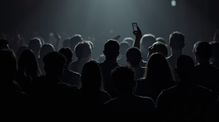 A crowd of people stands in a dimly lit venue facing a bright spotlight. Most faces are in shadow while one person holds up a phone, adding to the electric energy typical of live sports entertainment or concerts.