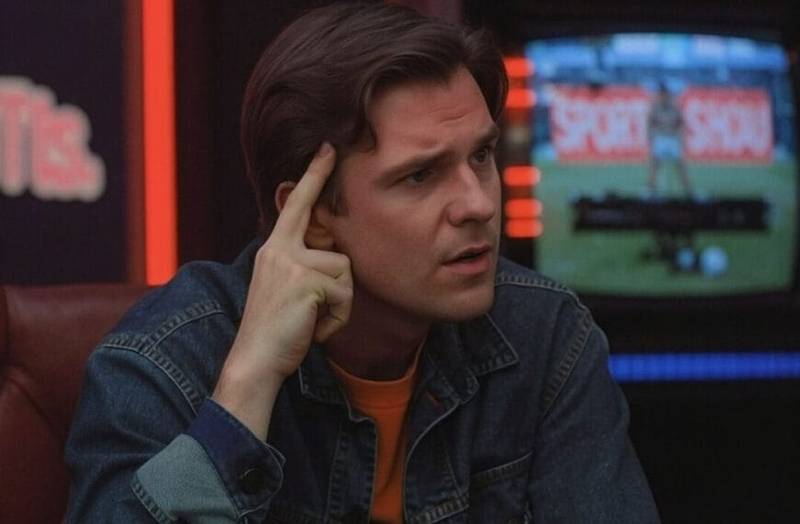 A young man with brown hair, wearing a denim jacket, sits indoors with his index finger pressed to his temple, looking thoughtful. Behind him, a blurred TV screen shows a sports broadcast featuring traditional sports commentary.
