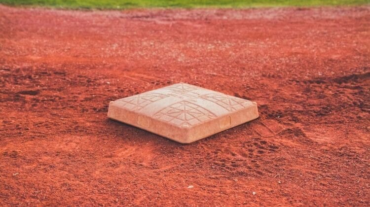 A close-up of a white baseball base sits on reddish-brown dirt, surrounded by a baseball infield. Reflecting Baseball Rules in the Dead Ball Era, the base’s textured surface and geometric patterns evoke classic field designs against green grass.