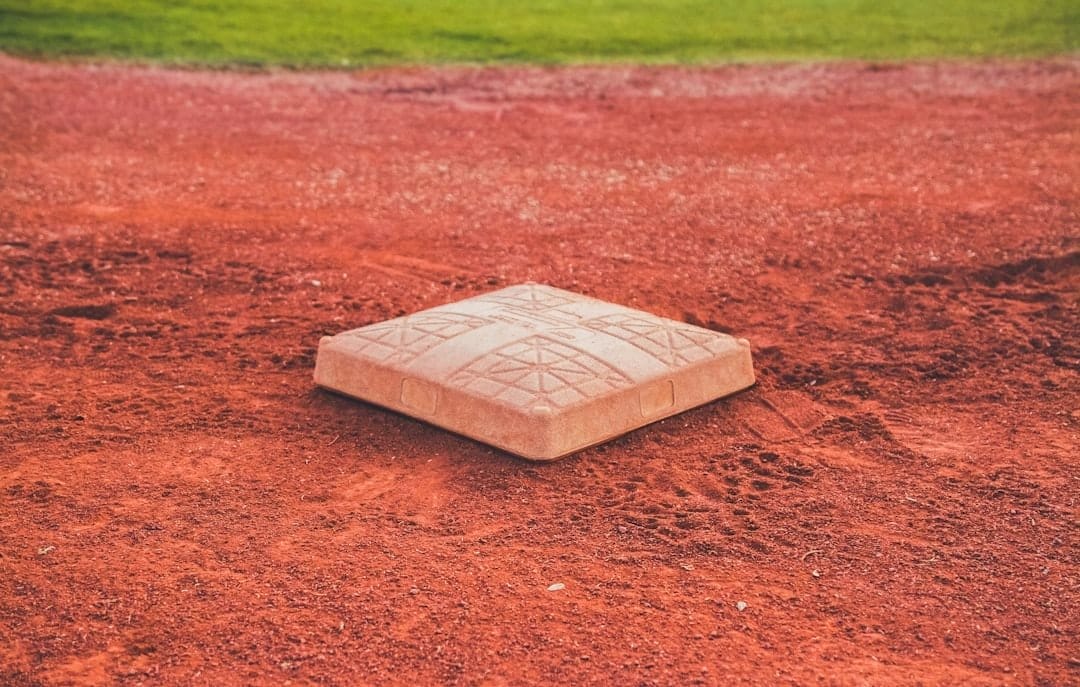 A close-up of a white baseball base sits on reddish-brown dirt, surrounded by a baseball infield. Reflecting Baseball Rules in the Dead Ball Era, the base’s textured surface and geometric patterns evoke classic field designs against green grass.