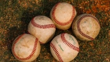 Five worn baseballs, reminiscent of the Dead Ball Era, with visible red stitching rest on a patch of grass and dirt. Warm lighting casts soft shadows, highlighting the textured surfaces and some faded green text on one of the balls.