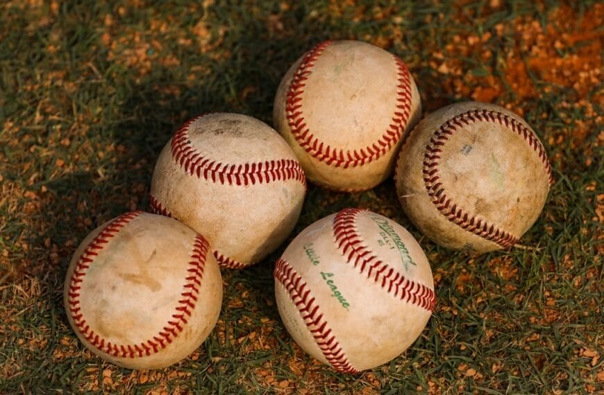 Five worn baseballs, reminiscent of the Dead Ball Era, with visible red stitching rest on a patch of grass and dirt. Warm lighting casts soft shadows, highlighting the textured surfaces and some faded green text on one of the balls.