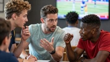Three men sit at a table having an animated discussion. Two are listening intently while the man in the middle gestures as he talks. A TV showing a sports event is blurred in the background. The setting appears casual and indoors.