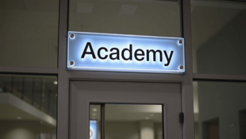 A glowing rectangular sign above a door reads "Academy" in bold black letters. The sign is mounted on a glass wall, and part of a modern, spacious interior with neutral colors is visible in the background.