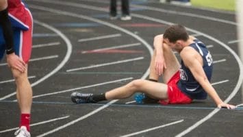 A male track athlete in a navy jersey and red shorts sits on the ground, holding his leg in apparent pain on a black running track. Another runner stands nearby bent over, while lanes and track lines are visible in the background.
