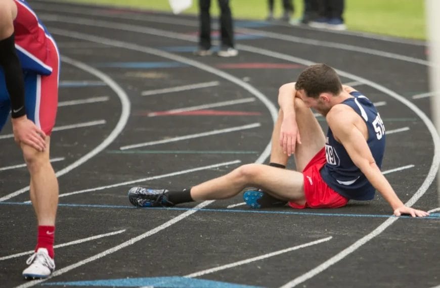 A male runner in a navy tank top and red shorts sits on a black outdoor track, leaning on one hand and clutching his leg in apparent pain. Sports argument strategies could be discussed as another fatigued runner stands nearby after the race.