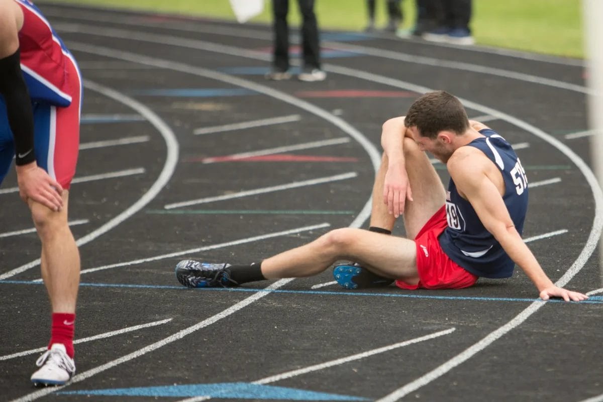 A male runner in a navy tank top and red shorts sits on a black outdoor track, leaning on one hand and clutching his leg in apparent pain. Sports argument strategies could be discussed as another fatigued runner stands nearby after the race.