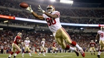 A San Francisco 49ers football player in a white jersey, number 80—often hailed as the greatest NFL player ever—dives mid-air with outstretched arms to catch a football in a brightly lit stadium filled with cheering fans and fellow players.