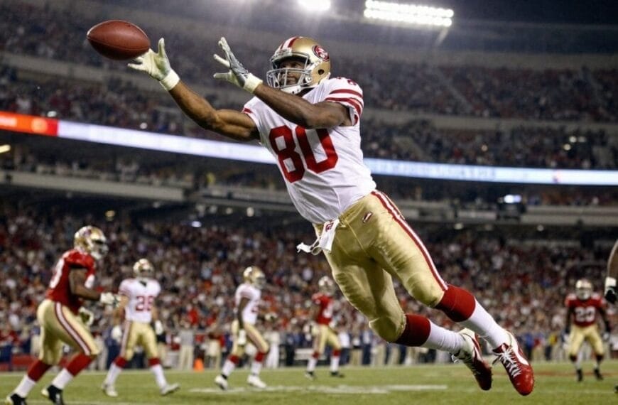 A San Francisco 49ers football player in a white jersey, number 80—often hailed as the greatest NFL player ever—dives mid-air with outstretched arms to catch a football in a brightly lit stadium filled with cheering fans and fellow players.