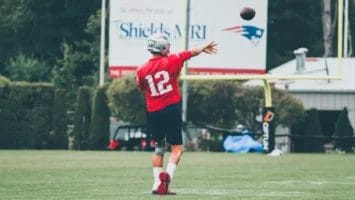 A football player in a red jersey with number 12 and helmet—often called the greatest NFL player ever—throws a football on a grassy field. Uprights, a billboard, and the Patriots logo appear in the background, with trees and equipment along the edge.