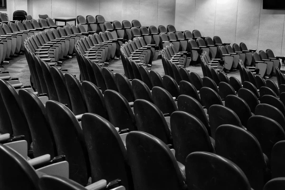 Black and white photo of an empty lecture hall with many curved rows of black chairs facing forward, armrests visible. The chairs are unoccupied, and the walls are light-colored with soft lighting creating shadows.