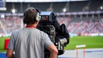 A camera operator wearing headphones films a sports event at a stadium, capturing moments often discussed in sports media criticism. The person stands behind a large broadcast camera, focused on the field, as a blurred crowd and green playing field fill the background.