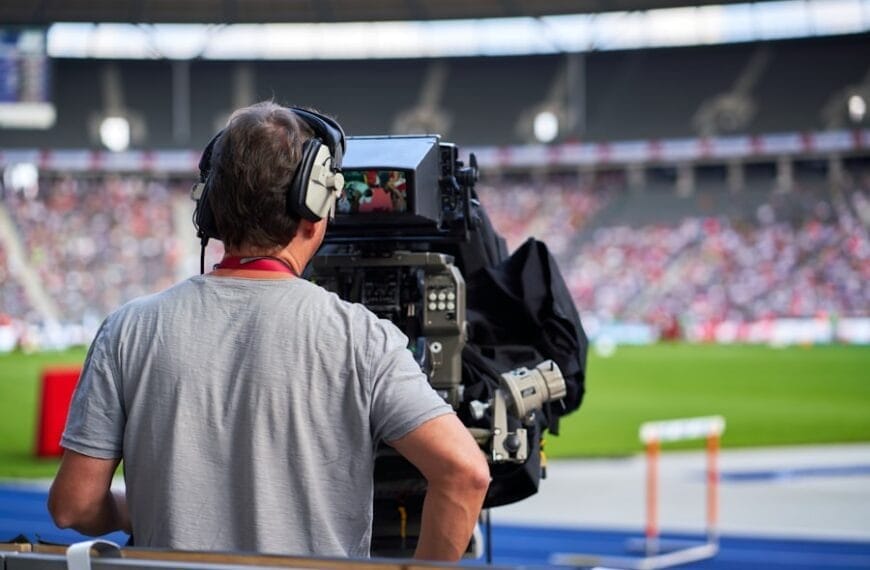 A camera operator wearing headphones films a sports event at a stadium, capturing moments often discussed in sports media criticism. The person stands behind a large broadcast camera, focused on the field, as a blurred crowd and green playing field fill the background.