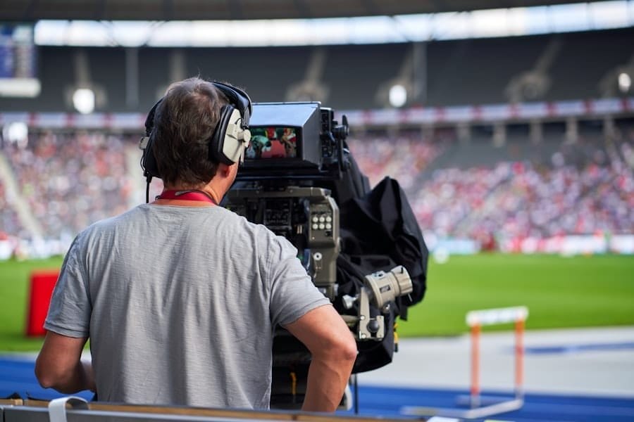 A camera operator wearing headphones films a sports event at a stadium, capturing moments often discussed in sports media criticism. The person stands behind a large broadcast camera, focused on the field, as a blurred crowd and green playing field fill the background.