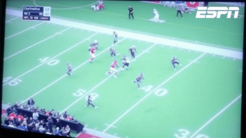 A televised football game screenshot showing a player in red running with the ball past the 40-yard line, pursued by several players in dark uniforms. The ESPN logo is in the upper right corner, and a score graphic is in the top left. Spectators are visible at the bottom.
