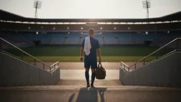 A soccer player in blue kit stands on stadium steps at sunset, facing an empty field. He holds a sports bag and white towel, with long shadows behind him—reminding us to respect sports and do not hate the sport players who give their all.