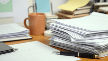 A cluttered desk with tall stacks of paper, a black pen on lined paper, a brown coffee mug, books, folders, and office supplies scattered in the background evokes the hectic pace of negotiating complex sports media deals.