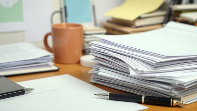 A cluttered desk with a thick stack of paper next to a coffee cup and a pen