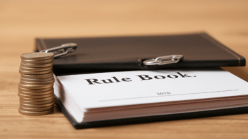 A stack of coins sits next to an open binder labeled "Rule Book" on a wooden surface, hinting at financial or regulatory matters—perhaps even highlighting salary cap structural flaws in the system.