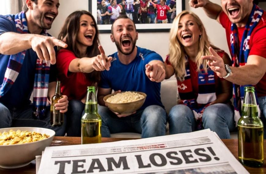 Five excited friends wearing team colors watch sports on TV, celebrating with snacks and drinks. A “CHAMPIONS!” photo hangs on the wall. On the table, a newspaper with “TEAM LOSES!” highlights confirmation bias in sports fans cheering anyway.